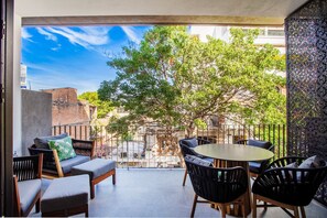 Relax on the balcony with a view of the tree-lined street, featuring a round table, comfortable seating, and a decorative wall.