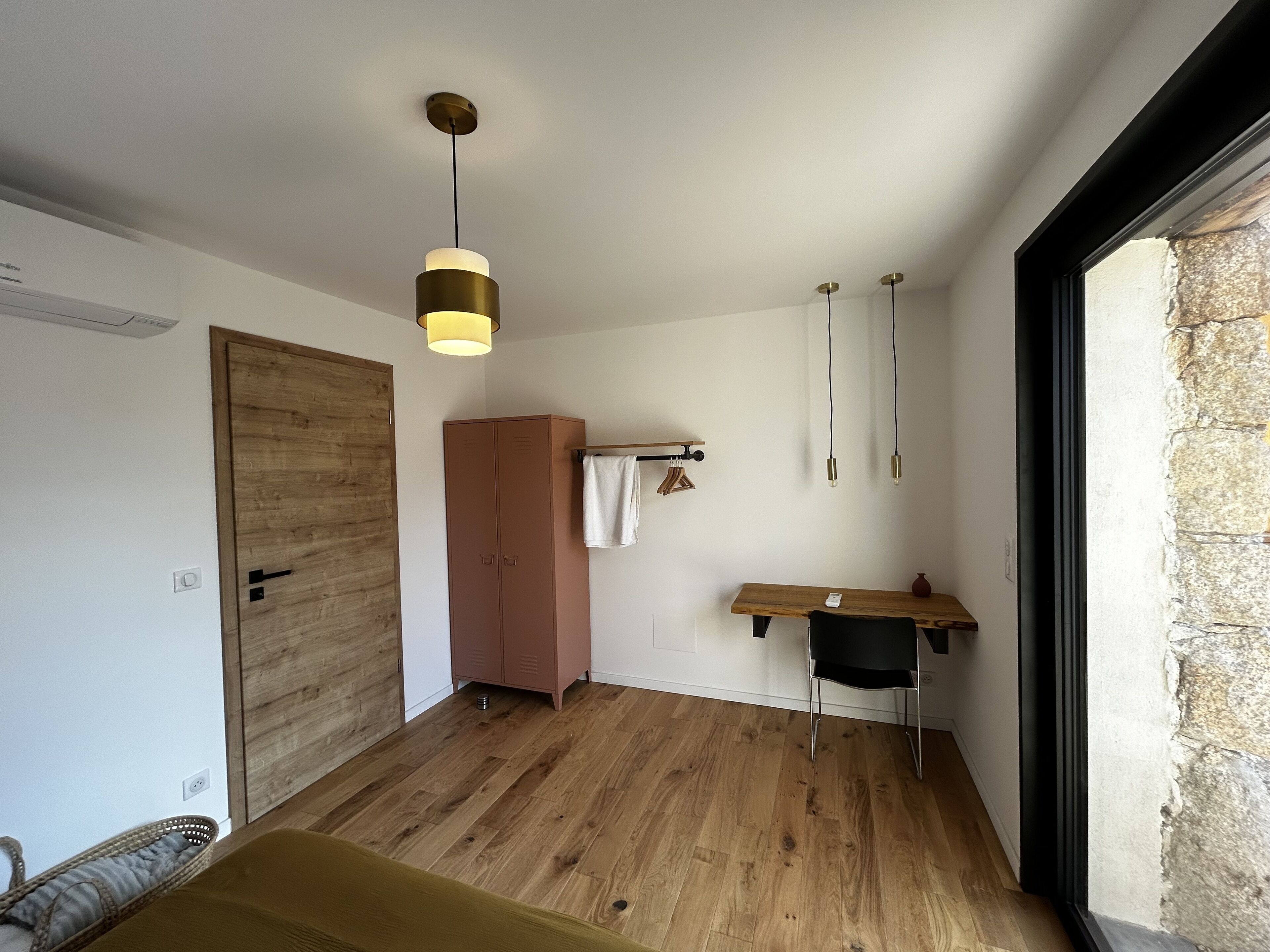 A room with a wooden wardrobe, a desk with a chair, a hanging light fixture, and a large window with stone wall.