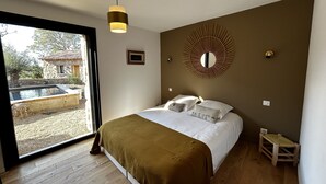 A bedroom with a bed, a small wooden stool, a round mirror, and a window with a view of a building and trees. - Maison de Type Bergerie , Proximité Porto Vecchio - Www.casarella.eu (Zonza)
