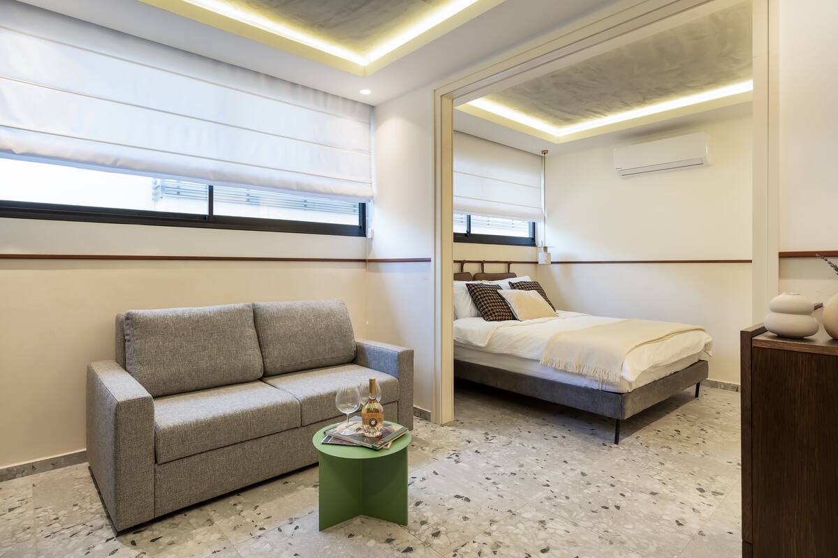A modern bedroom with a grey sofa, a green side table, a bed with white bedding, and a large mirror.