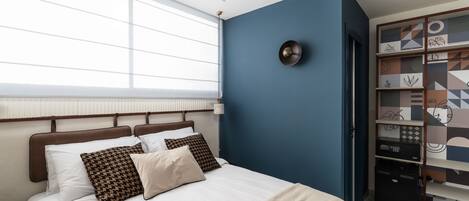 A bedroom with a bed, a nightstand, a wall-mounted lamp, and a window with blinds.