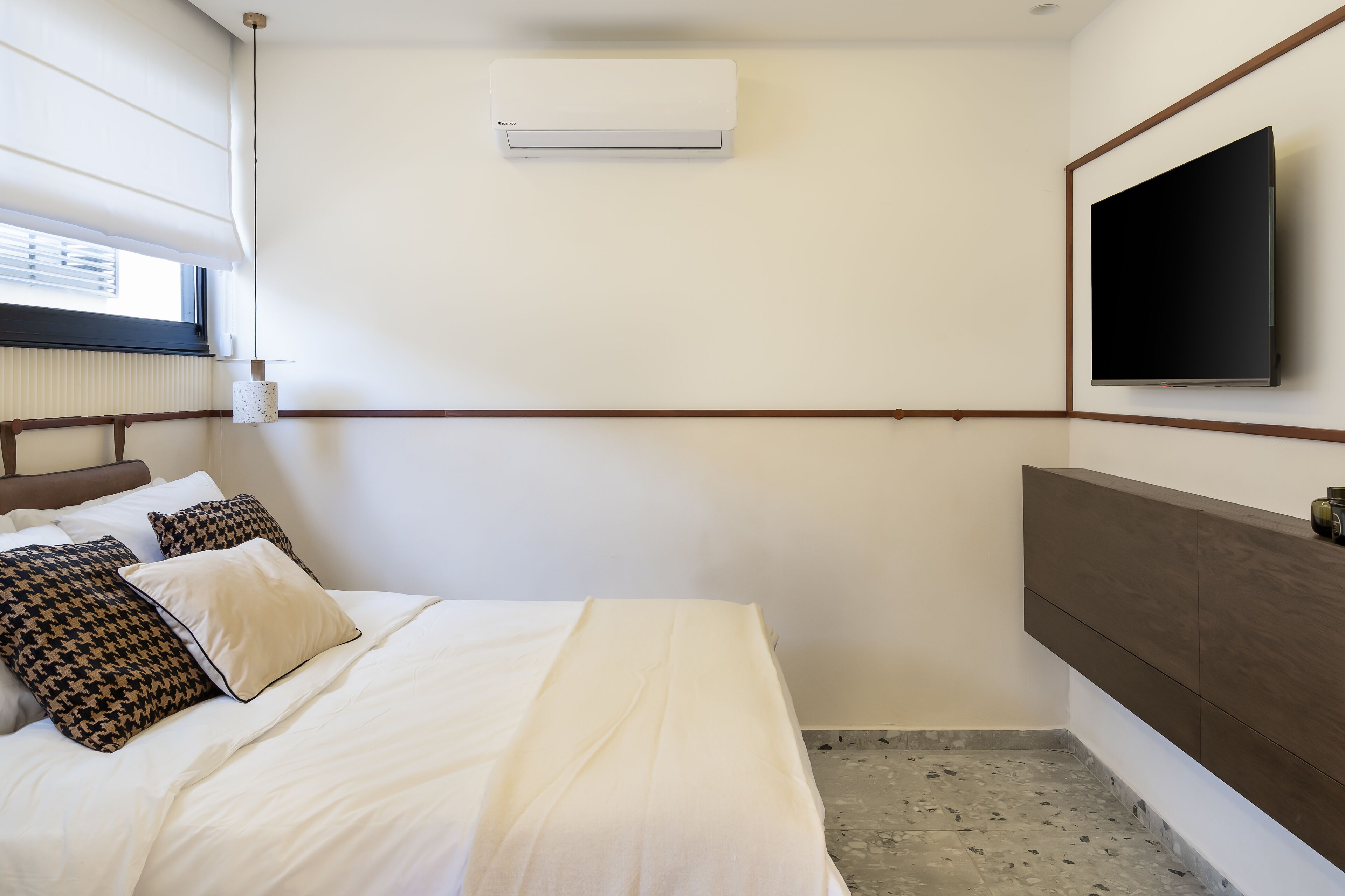 A hotel room with a bed, a TV mounted on the wall, an air conditioner, and a window with blinds.
