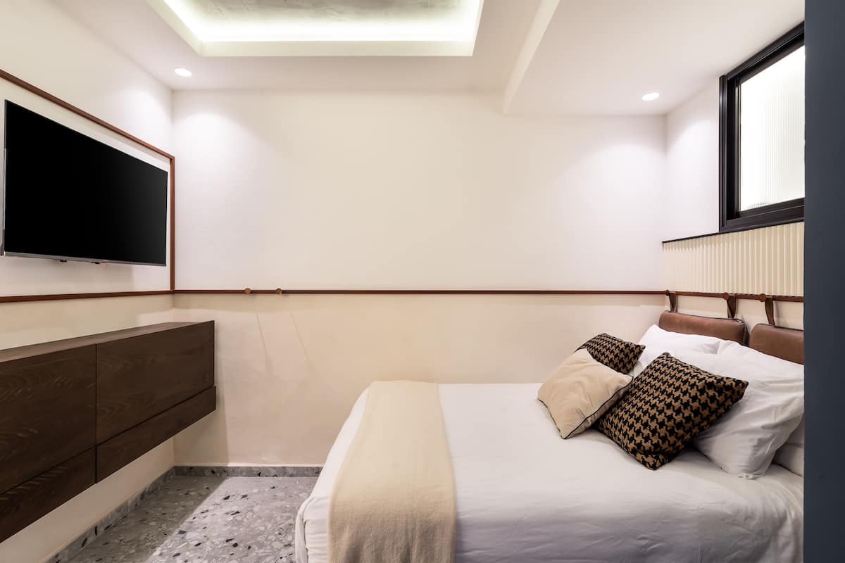 A modern bedroom with a large bed, a wooden bedside table, a flat-screen TV mounted on the wall, and a window with blinds.