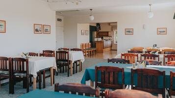 A dining area with multiple tables and chairs, a kitchen in the background, and framed pictures on the walls.