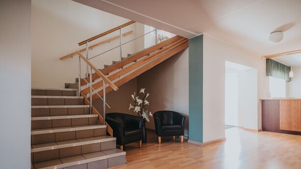 Una escalera de madera con barandillas de metal, que conduce a una zona de descanso con dos sillas negras y un jarrón de flores blancas.