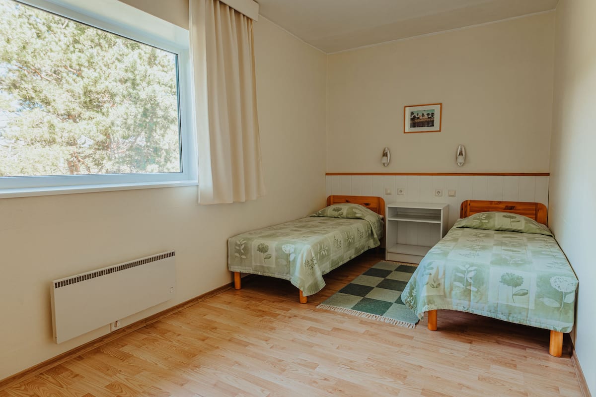Ein Zimmer mit zwei Einzelbetten, einem fensterbelaubten Fenster, einem Heizkörper und einem kleinen Schreibtisch mit Stuhl.