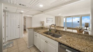A kitchen with granite countertops, a double sink, white cabinets, and a view of the ocean through large windows.