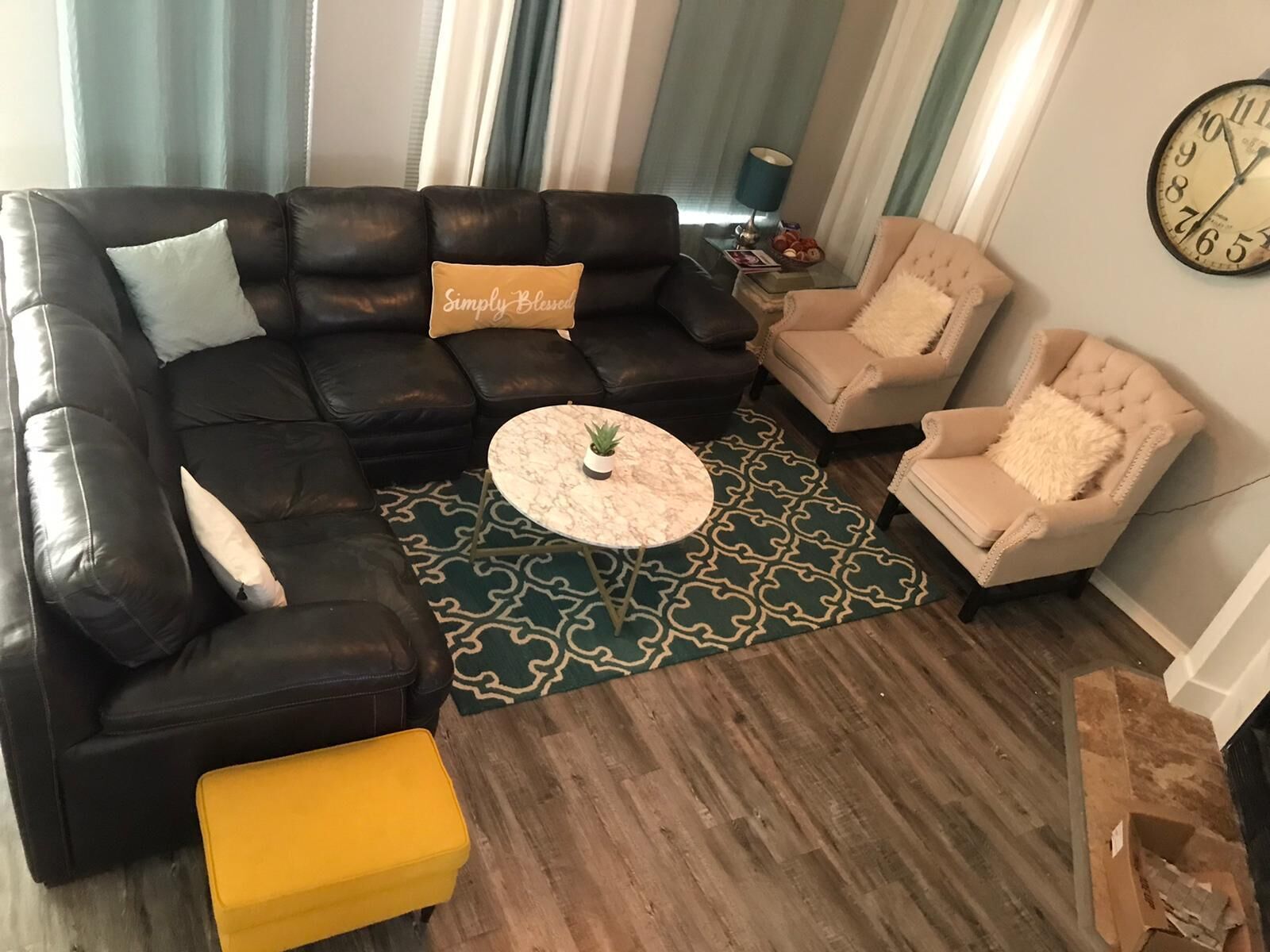 Un salon comprenant un canapé d’angle en cuir noir, un pouf jaune, un tapis à motifs, une table basse ronde et deux fauteuils beiges.