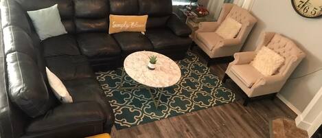Un salon comprenant un canapé d’angle en cuir noir, un pouf jaune, un tapis à motifs, une table basse ronde et deux fauteuils beiges.