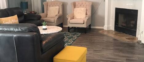 Un salón con un sofá de cuero negro, dos sillones beige, un reposapiés amarillo, un reloj redondo, una chimenea y un televisor sobre una repisa blanca.