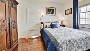 Ein Schlafzimmer mit einem Holzkleiderschrank, einem Bett mit gemusterten Betttextilien, einem Nachttisch mit einer Lampe und einem Fenster mit Vorhängen.