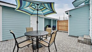 Eine Terrasse mit einem runden Tisch und Stühlen, einem gestreiften Sonnenschirm und hellblau verkleideten Gebäuden.