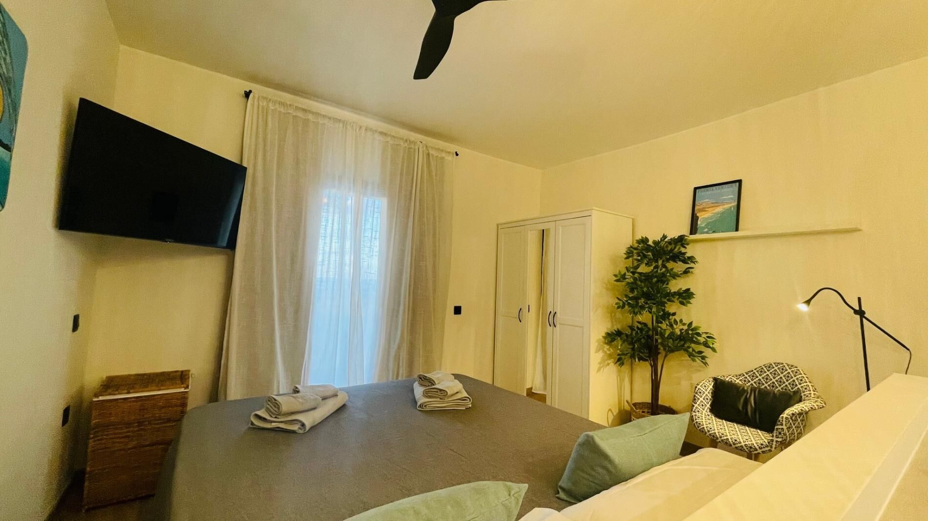 A hotel room with a bed, a flat-screen TV mounted on the wall, a wardrobe, a potted plant, and a bedside table with towels.