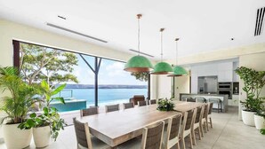 Een moderne eetruimte met een grote tafel en stoelen, grote ramen met uitzicht op zee, en een keuken op de achtergrond.