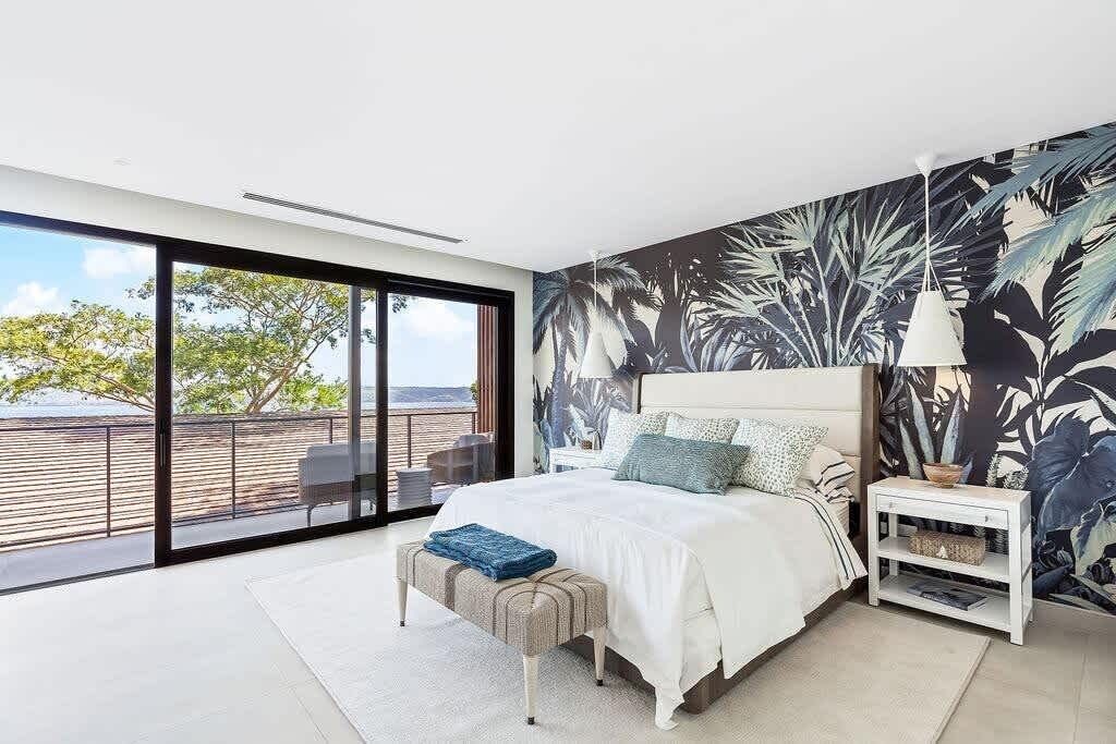 Une chambre dotée d’une grande porte-fenêtre coulissante donnant sur une terrasse extérieure, d’un lit avec une tête de lit blanche et d’un mur orné d’un motif de feuilles tropicales.