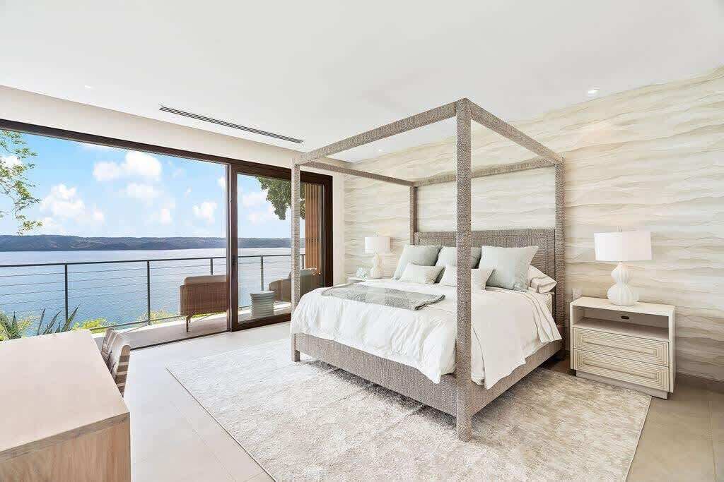 A modern bedroom with a four-poster bed, large windows, and a view of the sea.