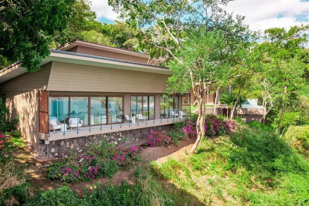 Una casa moderna con fachada de vidrio, mobiliario exterior y abundante vegetación.
