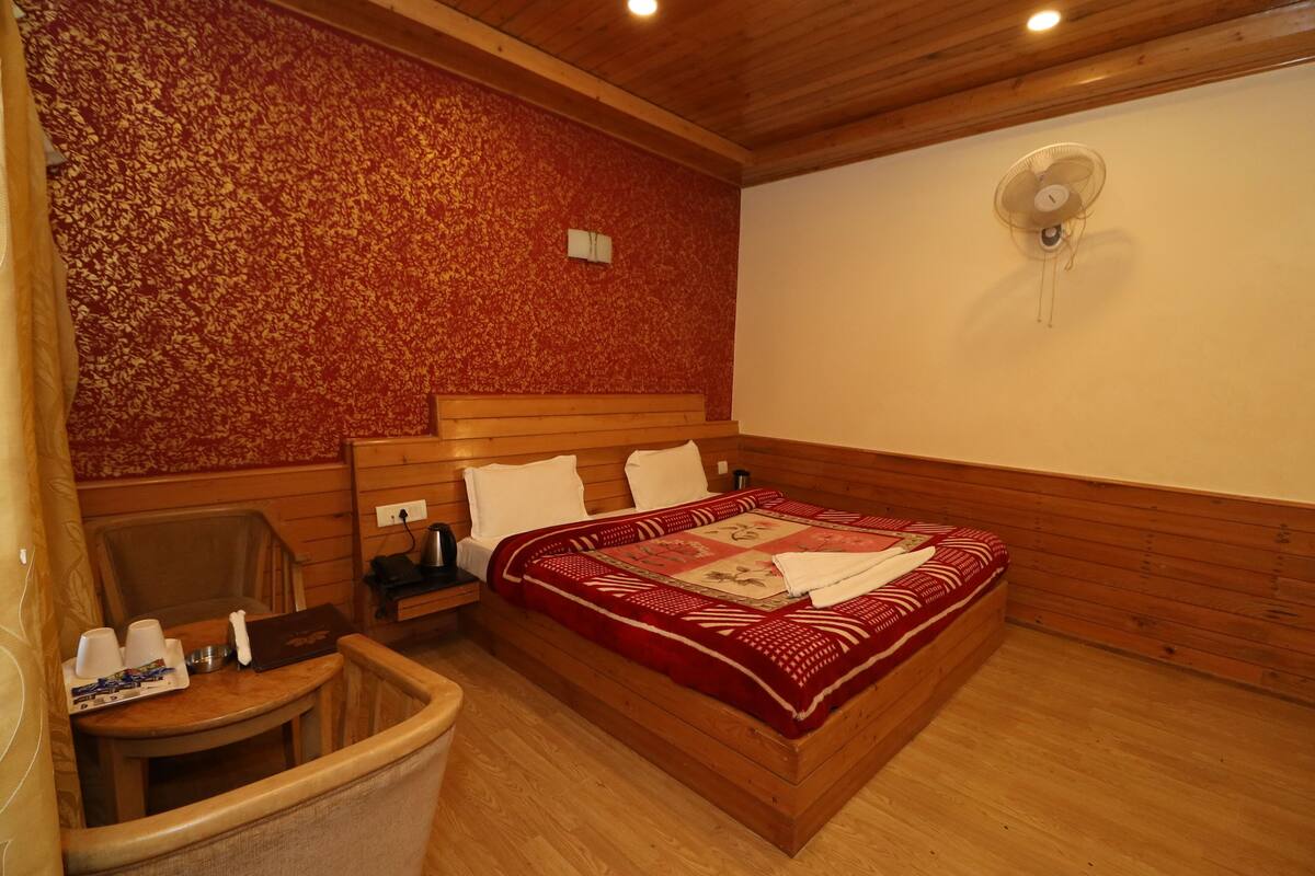A wooden-framed room with a bed, a chair, a small table, and a wall-mounted fan.