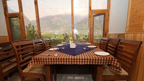 A dining area with a checkered tablecloth, wooden chairs, and a view of mountains. - Gezellig Inn - Golden Apple (Manali)