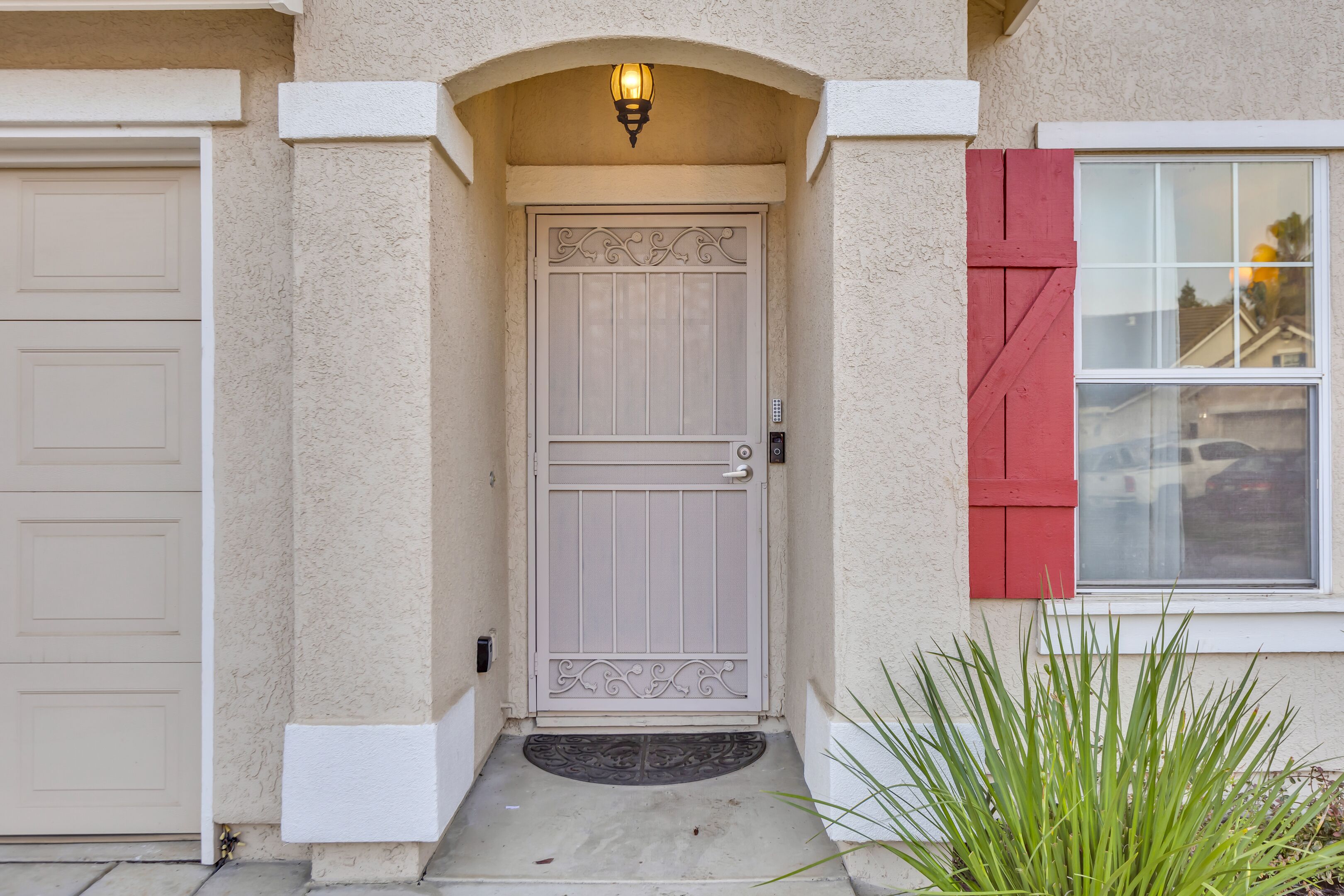 A house with a beige exterior, a white door with a decorative design, a red window shutter, and a small green plant in front.
