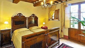 A traditional bedroom with a wooden bed, two bedside tables, a chandelier, and a window with curtains. - Estate in Monte Serra with Panoramic Views (Pisa)
