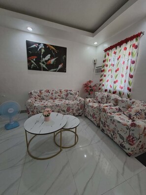A living room with a sofa set, a round coffee table, a fan, a painting, and a window with floral curtains.