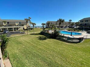 A pool area with a clear view of the ocean, surrounded by well-maintained grass and palm trees, with a building in the background.
