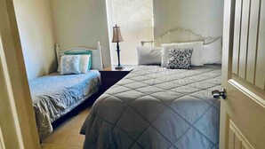 Quarto com duas camas, uma janela com persianas, um abajur e uma porta.