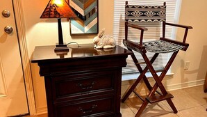 Uma mesa de cabeceira de madeira com gavetas e uma luminária, uma cadeira estampada e uma estatueta decorativa de coelho.
