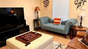Uma sala de estar com televisão, um sofá azul, uma mesa de centro com jogo de tabuleiro e arte decorativa nas paredes.