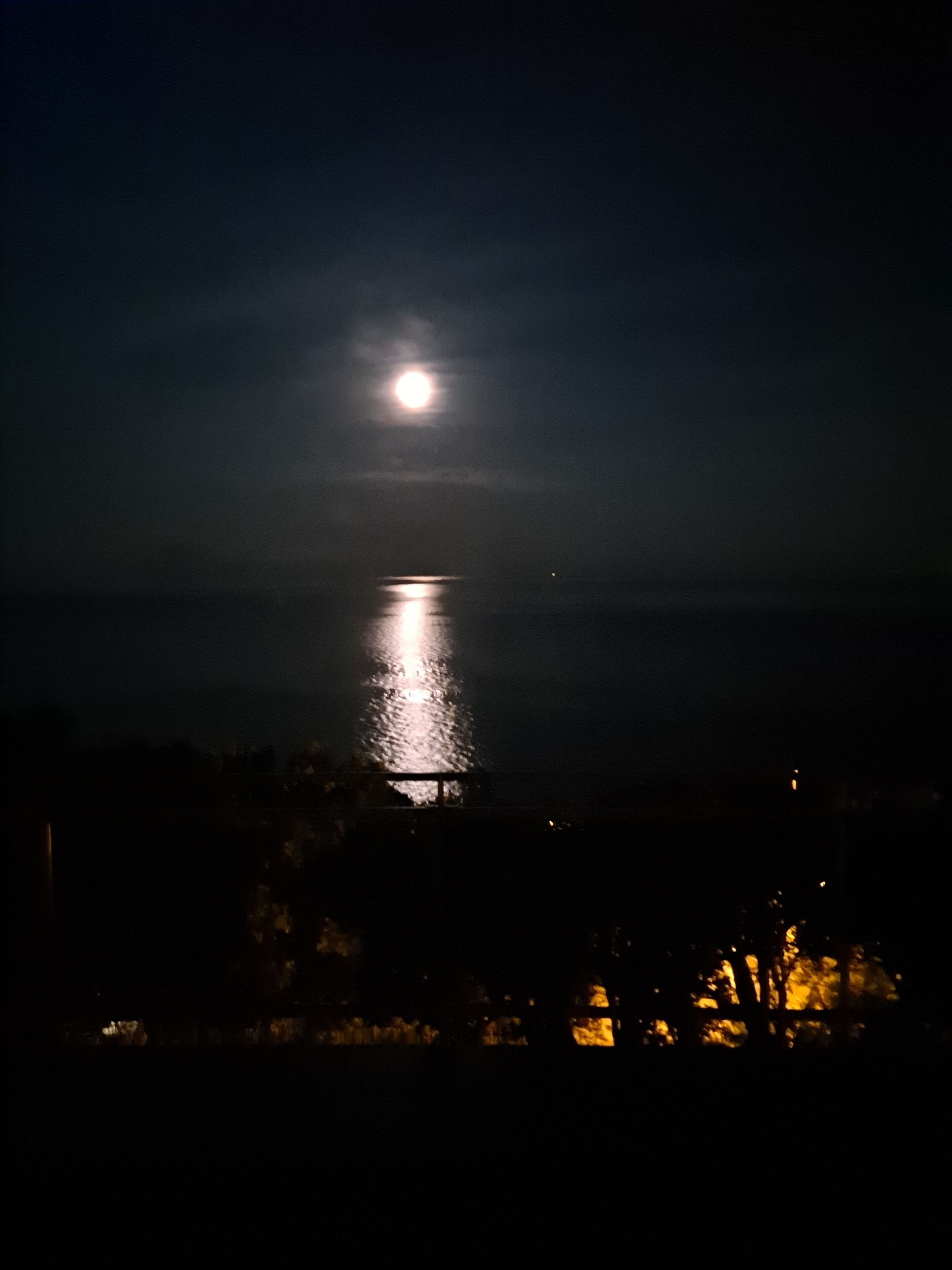 Une scène nocturne où l’on aperçoit le reflet de la lune sur une étendue d’eau, le tout étant entouré d’un paysage sombre parsemé de quelques lumières lointaines.