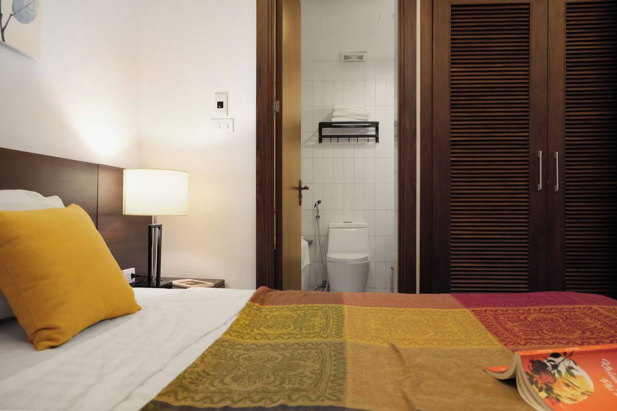 A hotel room with a bed, a nightstand, a lamp, a bathroom visible through an open door, and a wooden wardrobe.