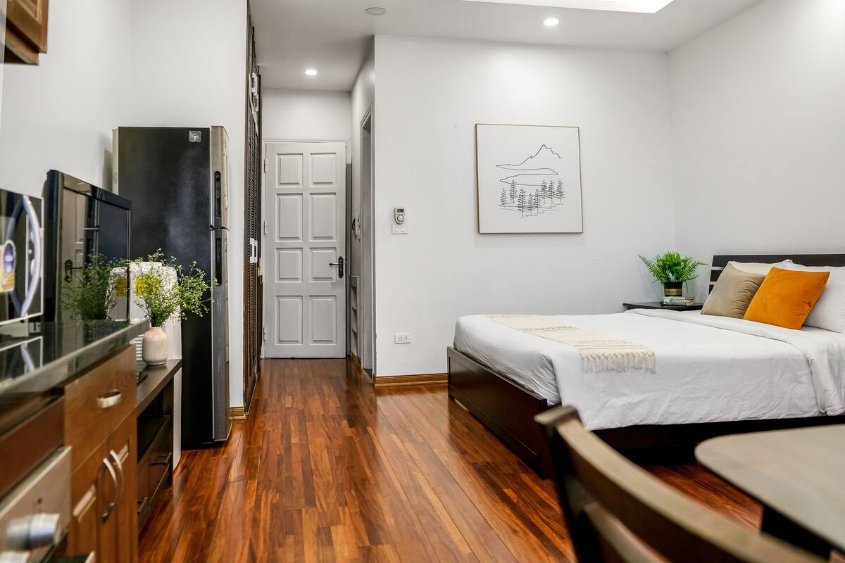 A modern hotel room with a wooden floor, a bed with white bedding, a small table, and a TV on a wooden cabinet.