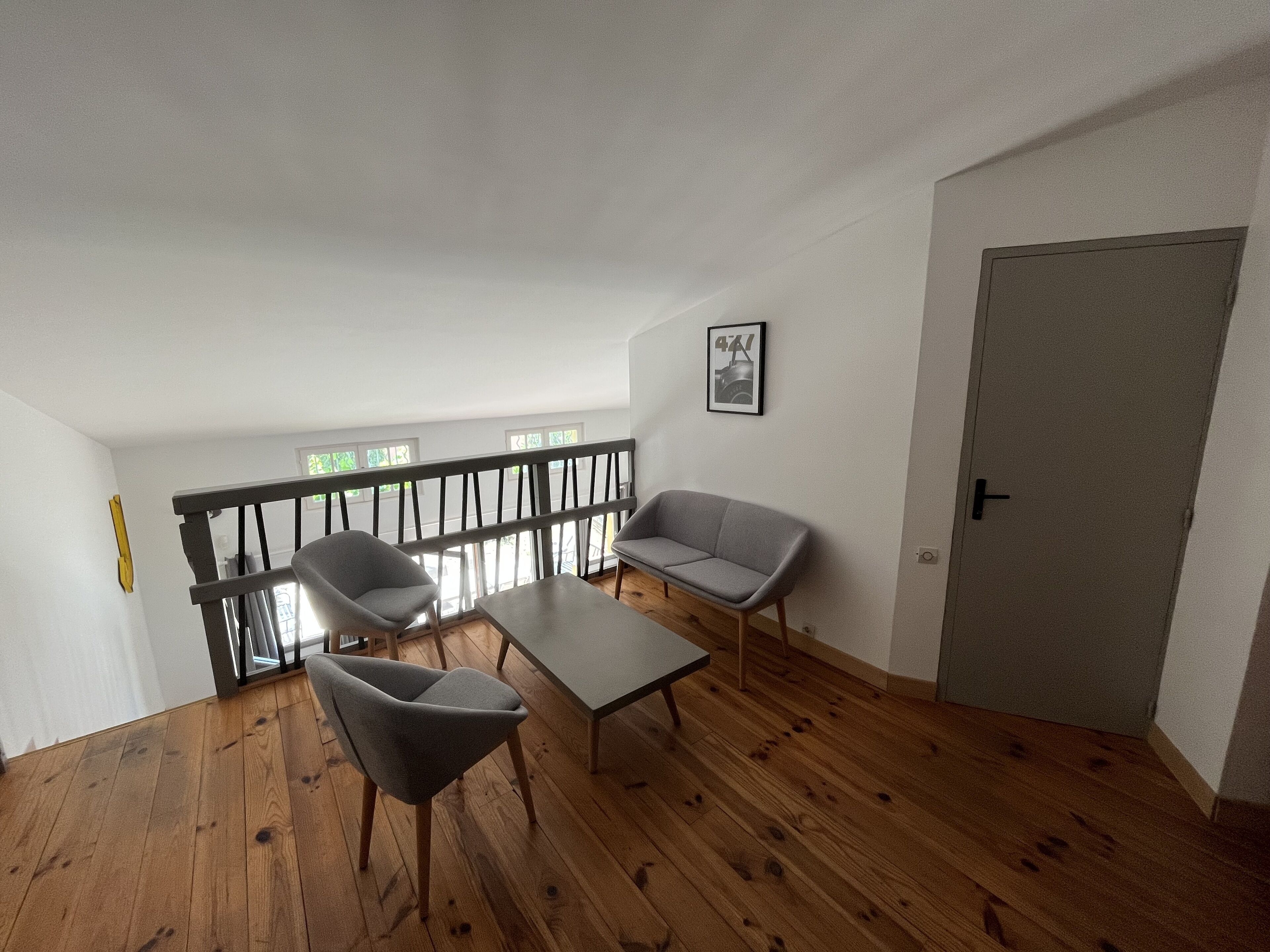Um quarto com piso de madeira, duas cadeiras cinzas, um sofá cinza, uma mesa pequena, uma porta e uma janela com vista para árvores.