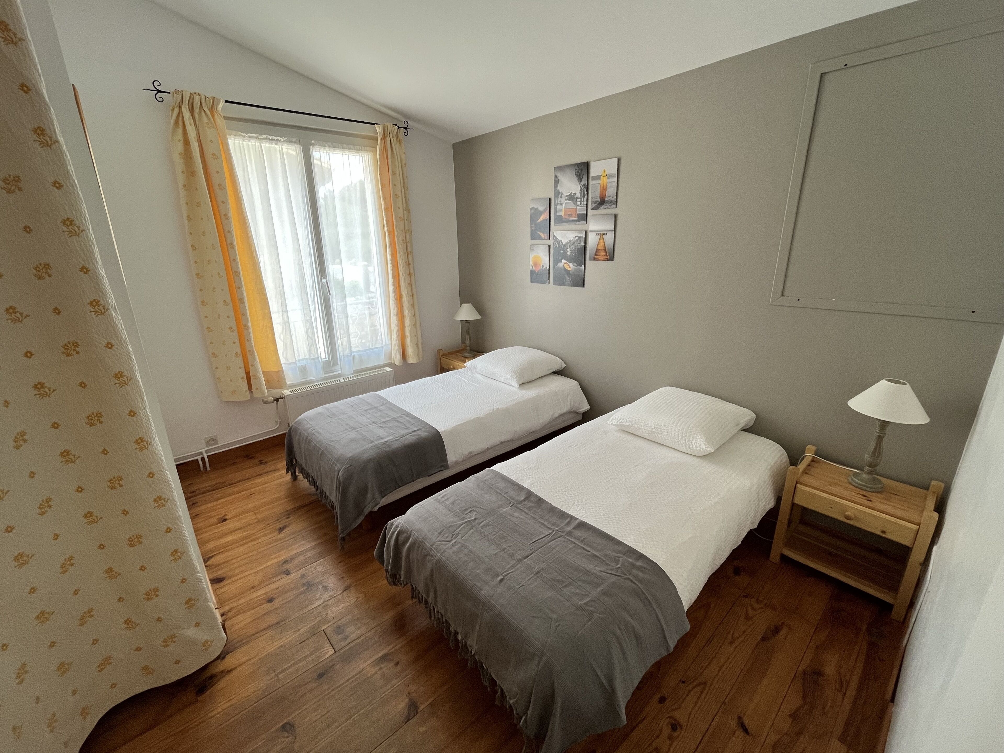 A bedroom with two single beds, a window with curtains, a bedside table with a lamp, and a wall with framed pictures.