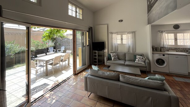 A spacious living area with a sliding glass door leading to a patio, a grey sofa, a flat-screen TV, and a kitchen with a washing machine. - Shelby - Gîte Atypique 6 Personnes à 5mn D'uzès Style Loft Ambiance Automobile (Saint-Quentin-la-Poterie)