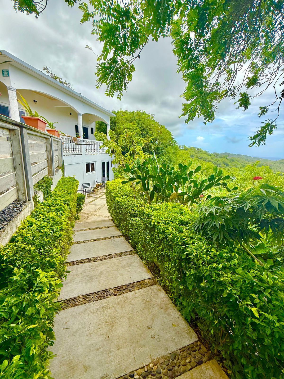 A pathway leading to a white building with a balcony, surrounded by lush greenery and a mountainous backdrop.