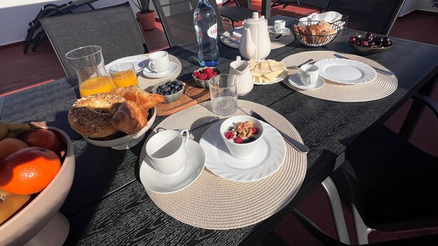 Une table de petit-déjeuner avec des croissants, des fruits et des boissons, sur une terrasse offrant une vue sur la ville et la mer.