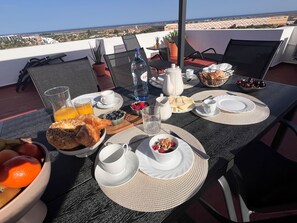 Une table de petit-déjeuner avec des croissants, des fruits et des boissons, sur une terrasse offrant une vue sur la ville et la mer.