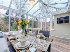 A conservatory with a dining table set for four, surrounded by large windows offering a view of trees and hills.