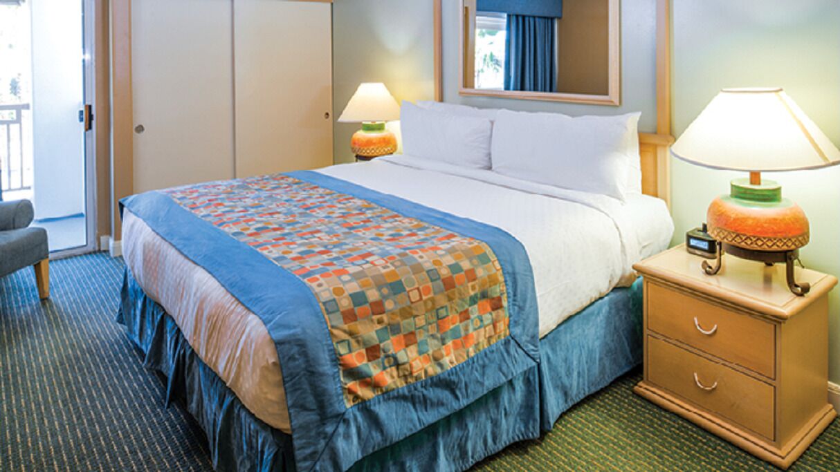 Una habitación de hotel con una cama, una mesita de noche con lámpara, una silla y una puerta que da a un balcón.