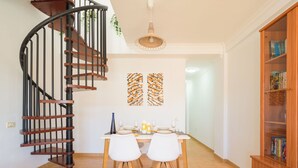 Un coin repas avec une table en bois, deux chaises blanches, un escalier en colimaçon et une bibliothèque.