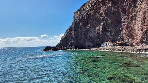 Un paysage côtier avec une eau turquoise et limpide, une falaise rocheuse et un petit bâtiment doté d’un toit rouge, situé près du rivage.