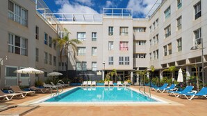 Une piscine entourée de chaises longues et de parasols, le tout bordé d’un bâtiment doté de nombreuses fenêtres et de balcons.