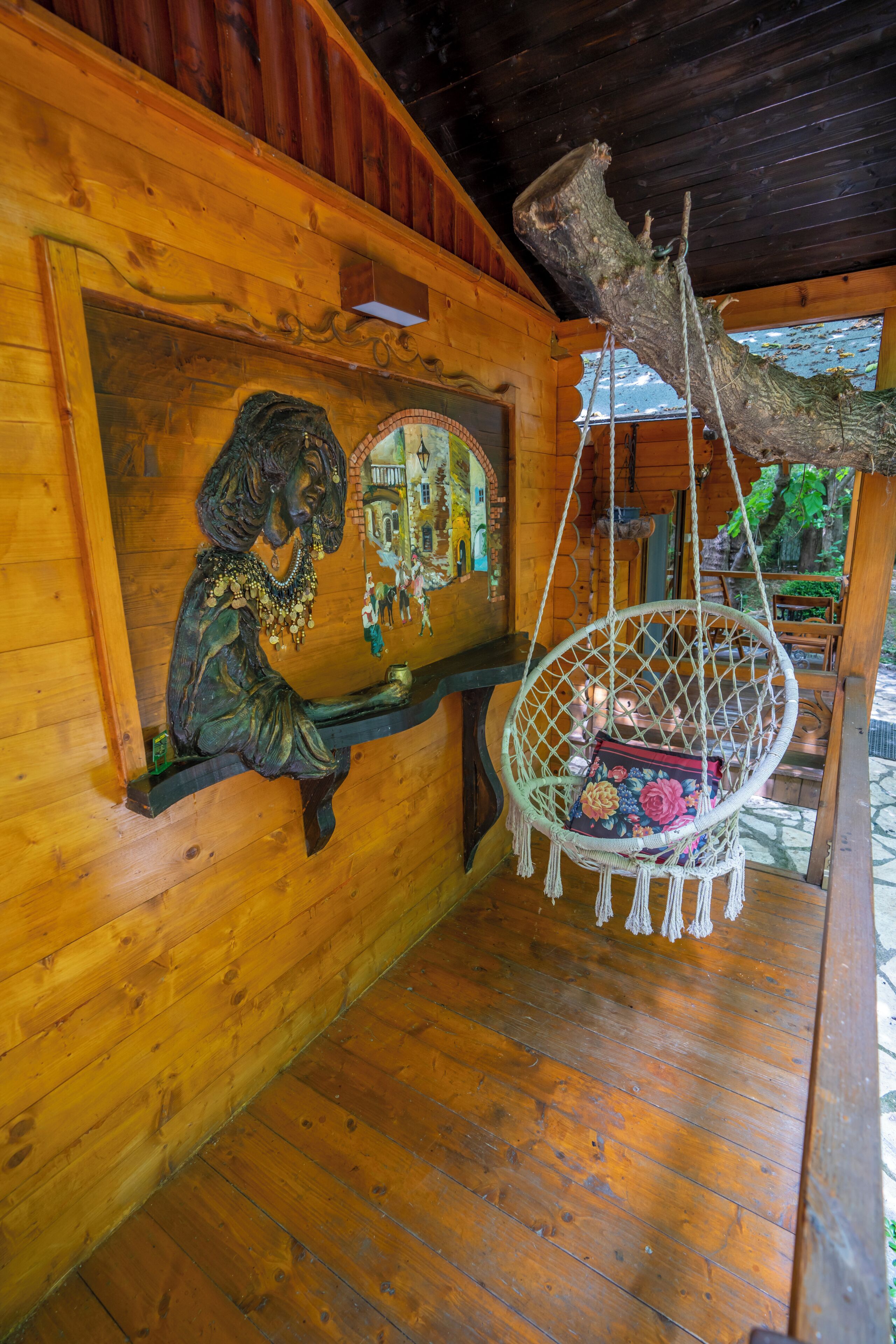 Interior de una cabaña de madera con un mural, una silla hamaca y techo rústico.