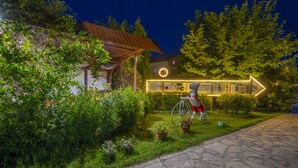 A stone pathway leading to a building with a metal roof, surrounded by lush greenery and a directional sign. - Hotel Legjenda (Shkodër)