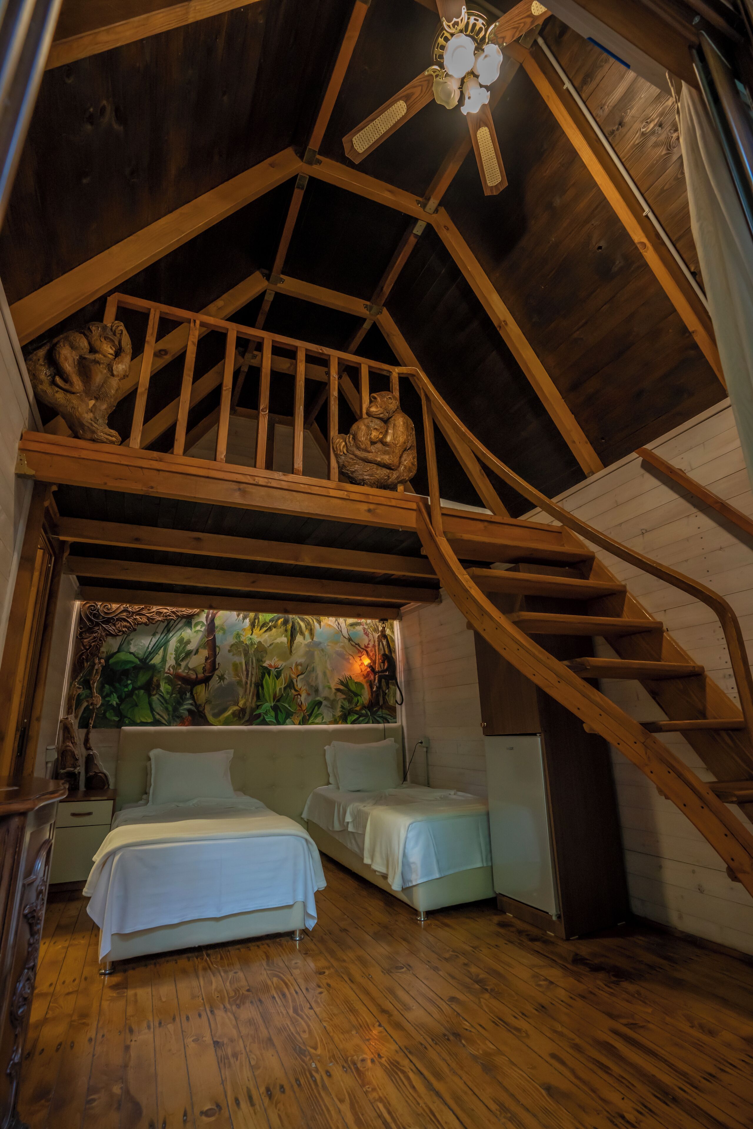 A bedroom with a sloped ceiling, wooden beams, a bed, a sofa, and a view of a forest through a window.