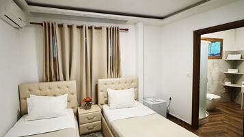 Habitación de hotel con dos camas, una mesita de noche pequeña, un armario, una ventana con cortinas, un baño visible a través de una puerta abierta y piso de madera.
