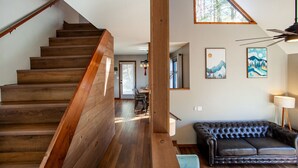 Una escalera de madera conduce a un comedor con vistas al exterior. En primer plano, un sofá de cuero negro y una lámpara.