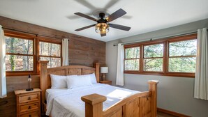 Dormitorio con estructura de madera, cama, mesitas de noche, ventilador de techo y ventanas amplias.
